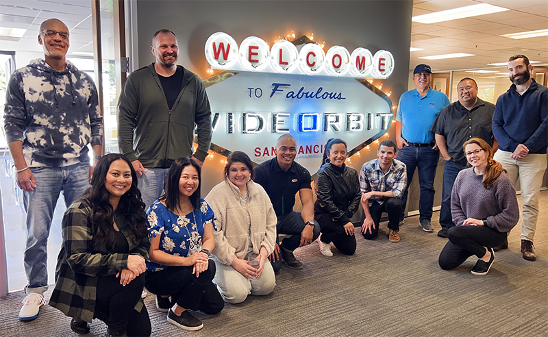 Team of WideOrbit employees at San Francisco Headquarters near the Vegas-inspired light up sign that reads "Welcome to the fabulous WideOrbit"